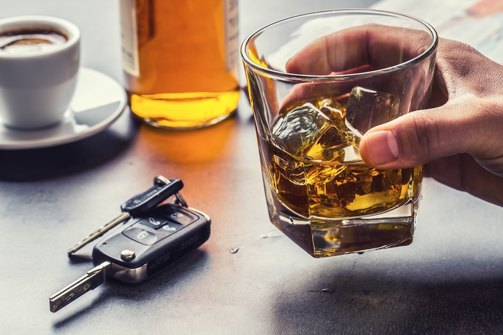 A hand holding a glass of whiskey with ice beside a car key, bottle, and cup of coffee on a table, suggesting the dangers of drinking and driving.