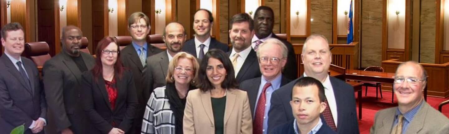 A group of thirteen professionally dressed people pose and smile together in a formal courtroom setting with wooden benches and judicial seating visible in the background.