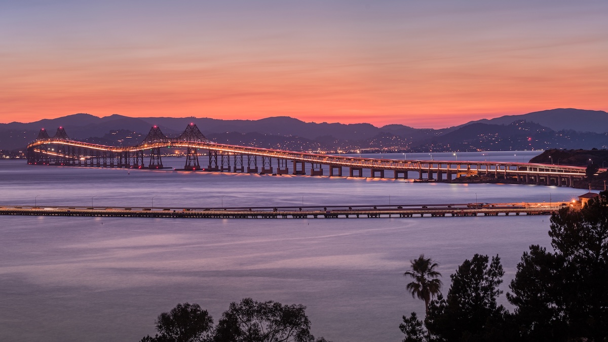 A long, illuminated bridge curves over a calm bay at sunset, with pink and orange hues in the sky and mountains in the background. Silhouettes of trees and a palm are visible in the foreground.