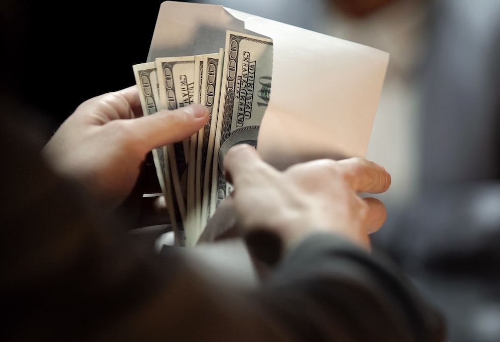 A person holding several hundred-dollar bills and placing them into a white envelope, with a blurred background.