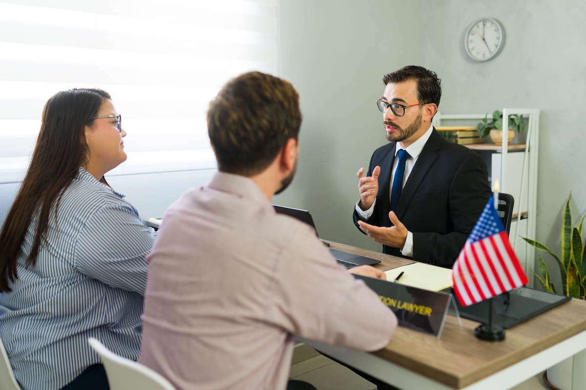 A man in a suit speaks to a seated couple in an office. A small American flag and a sign reading immigration lawyer are on the desk. A clock and bookshelf are in the background.
