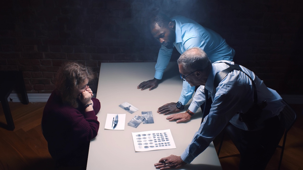 Two men, possibly detectives, stand over a table questioning a seated woman. The table has documents, fingerprints, and small plastic bags, suggesting a police interrogation scene.