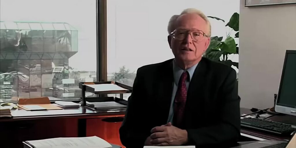 An older man in a suit and glasses sits at a desk in an office.